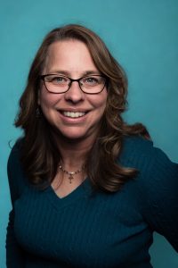 Smiling woman with long wavy brown hair wearing glasses, a teal sweater, and a cross necklace, in front of a blue background.