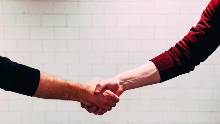 Two people shaking hands in front of a white tile wall, symbolizing connection and trust.