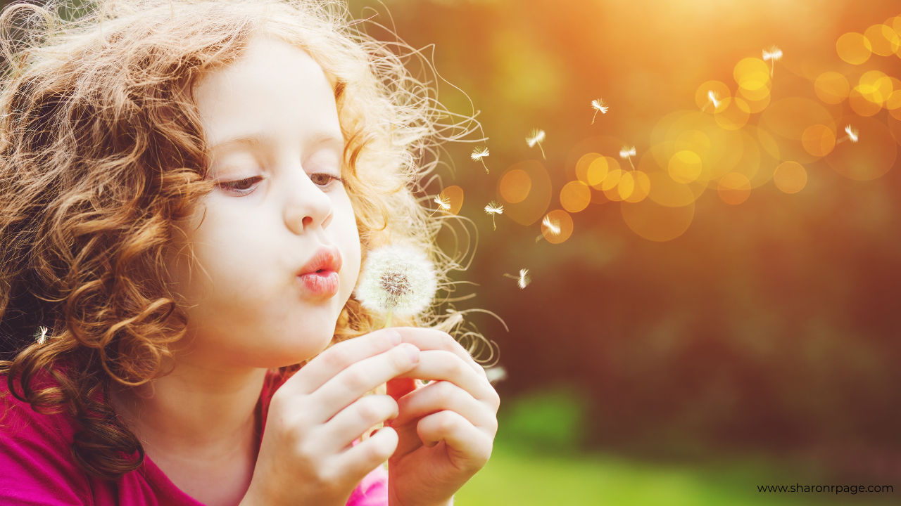 Young girl blowing on a dandelion puff in warm evening light, with seeds drifting through the air.
