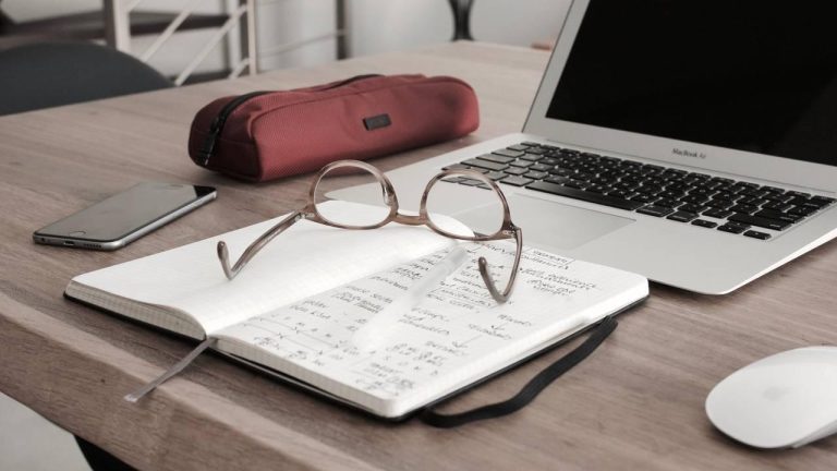 Open journal with handwritten notes, eyeglasses resting on the page, beside a laptop and coffee cup on a wooden desk—suggesting reflection, planning, and intentional work.