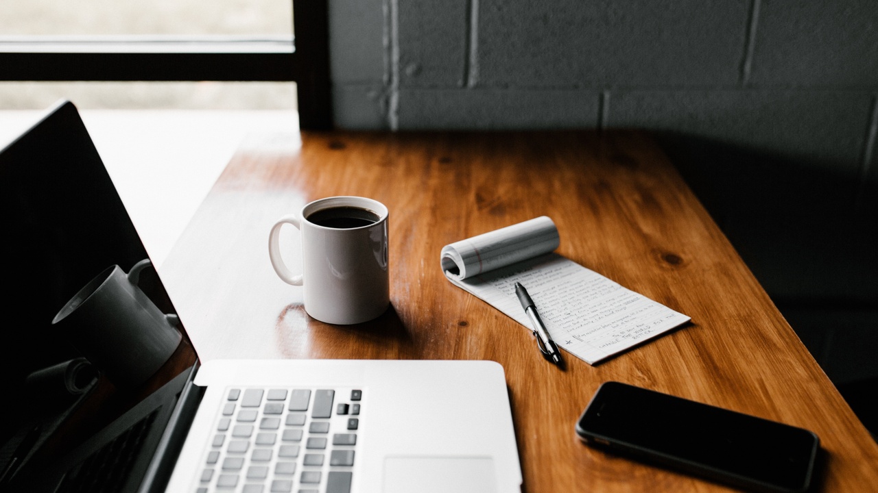A wooden desk with a laptop, a white coffee mug, a spiral notepad with handwritten notes, a pen, and a smartphone—suggesting a quiet, productive workspace.