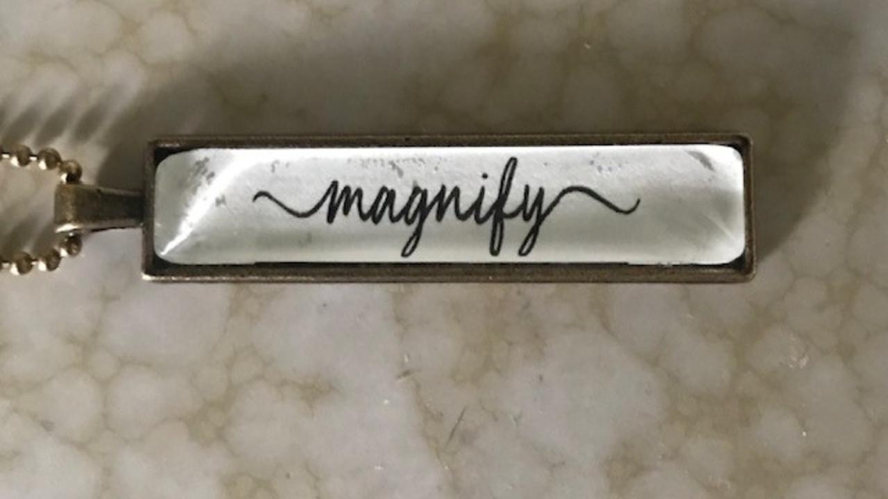 Close-up of a bronze pendant necklace with the word “magnify” written in cursive on a white background.